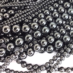 May include: A close-up of a collection of black hematite beads in various sizes. The beads are smooth and shiny, with a metallic sheen. Some beads are round, while others are slightly oval.