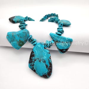 May include: A turquoise necklace featuring large, irregularly shaped stones. The necklace has a striking blue color with dark brown veining, and is strung together with smaller, rounded turquoise beads. The design is set against a white background.