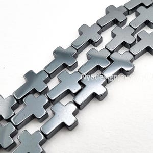 May include: Close-up of several strands of gray cross-shaped beads. The beads are made of a smooth, reflective material and are arranged in parallel rows. The crosses have a simple, modern design.