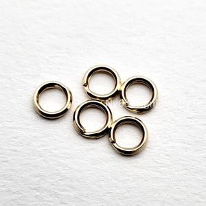 May include: Five gold-colored jump rings are arranged on a white surface. The rings are circular with a small opening, suggesting they are for jewelry making or crafts. The rings have a shiny, metallic finish.