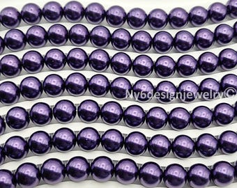 May include: Close-up of several strands of dark purple, glossy beads. The beads are round and evenly spaced, creating a uniform pattern. The background is a white, curved surface. The beads are likely for jewelry making.