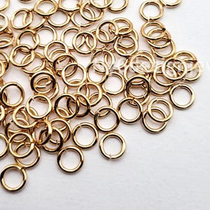 May include: A collection of small, gold-colored jump rings scattered on a white surface. The rings are circular with a small opening, suggesting they are for jewelry making or crafts. The lighting highlights the metallic sheen of the rings.