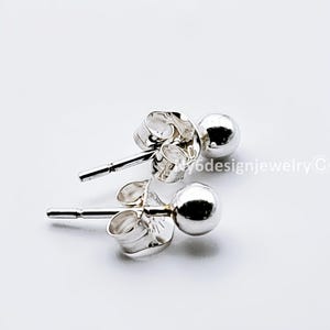 May include: A pair of small, shiny silver ball stud earrings. The earrings have a post and butterfly backing. The earrings are displayed on a white background. The earrings are a simple, classic design.