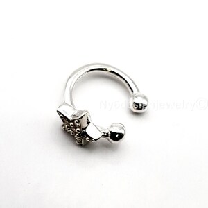 May include: A silver-colored, horseshoe-shaped ear cuff. The cuff features a decorative rectangular element with small, raised details and two rounded ball accents. The text "Ny6designjewelry©" is visible.