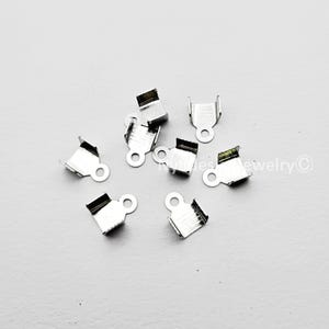 May include: A collection of silver-colored crimp end findings for jewelry making. Each piece has a rectangular shape with a loop for attaching to other components. The findings are designed to secure the ends of cords or ribbons.