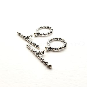 May include: Close-up of silver-toned jewelry clasps. The clasps include two T-bars and two rings with a twisted rope design. Each ring has a small, clear gem. The clasps are on a white background.