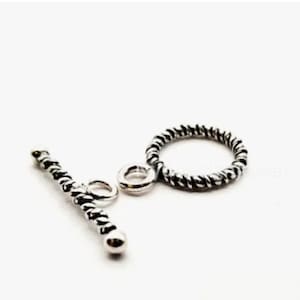 May include: A silver-toned toggle clasp for jewelry making, consisting of a circular ring and a T-shaped bar. Both components feature a textured, twisted rope-like pattern with darkened recessed areas to highlight the metallic detail.