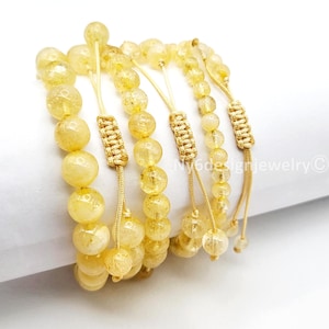 May include: Several strands of yellow gemstone bead bracelets. Each bracelet features round, translucent beads and gold-colored macrame knots. The bracelets are displayed on a white surface, with the jewelry arranged in a layered composition.