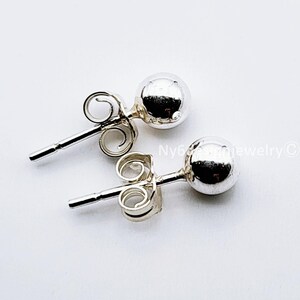 May include: A pair of shiny, silver-colored ball stud earrings. Each earring features a small, round ball attached to a post with a butterfly backing. The earrings are displayed on a white background.