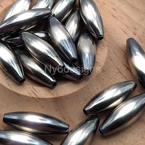 May include: A close-up of a pile of silver-colored, oval-shaped beads. The beads are smooth and shiny, and they have a slightly iridescent finish.