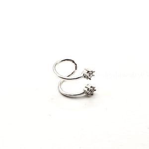 May include: A silver-colored, spiral-shaped ear cuff with two square, clear gemstones. The ear cuff is made of a shiny metal and is designed to wrap around the ear. The jewelry is set against a white background.