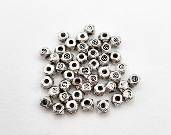 May include: A collection of small, silver-colored metal beads. Each bead has a hexagonal shape with a textured surface and a circular hole. The beads are scattered on a white surface.