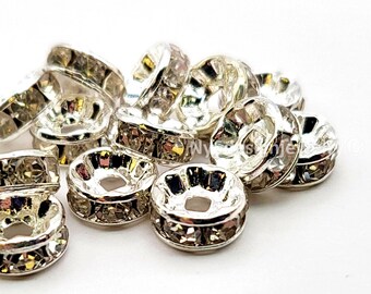 May include: Close-up of several silver-colored spacer beads with a faceted, crystal-like appearance. The beads have a circular shape with a large central hole and are designed for jewelry making. The beads are scattered on a white surface.