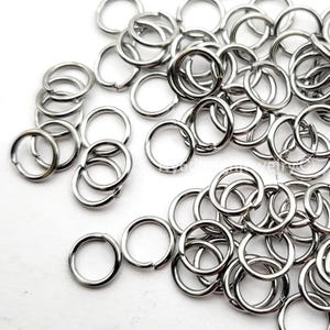 May include: A collection of shiny, silver-colored split rings, scattered across a white surface. These small, circular metal rings are open at one point, designed for connecting jewelry components or other craft projects. The rings are made of a reflective metal.