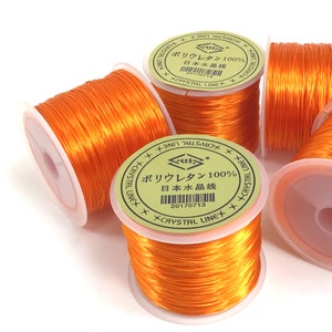 May include: Several spools of bright orange elastic cord are displayed against a white background. The spools have white centers and are labeled with Japanese text and the words "CRYSTAL LINE". The cord appears to be made of a shiny, flexible material.