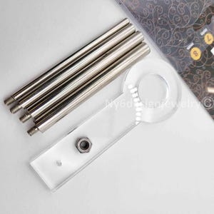May include: A collection of silver-colored metal rods with threaded ends, a clear acrylic piece with a circular design, and a hexagonal nut. The items are arranged on a light-colored surface. The text "Ny6d design jewelry" is visible.