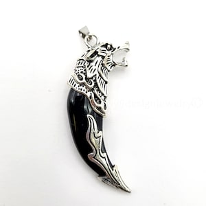 May include: A silver-toned wolf head pendant with a black, tooth-shaped centerpiece. The wolf head features detailed fur and an open mouth. The pendant is designed to be worn as a necklace.