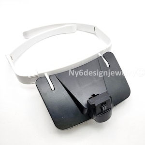 May include: A white headband with a black, adjustable magnifier. The magnifier has a rectangular shape with a small, adjustable knob. The headband is made of a lightweight material.