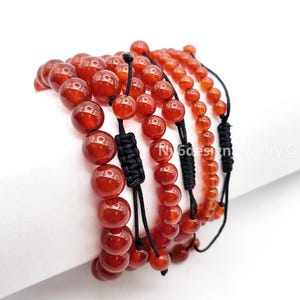 May include: Several red carnelian bead bracelets with black adjustable cords. The bracelets feature various sizes of polished, round beads. The bracelets are displayed against a white background. The jewelry is from N6designjewelry.