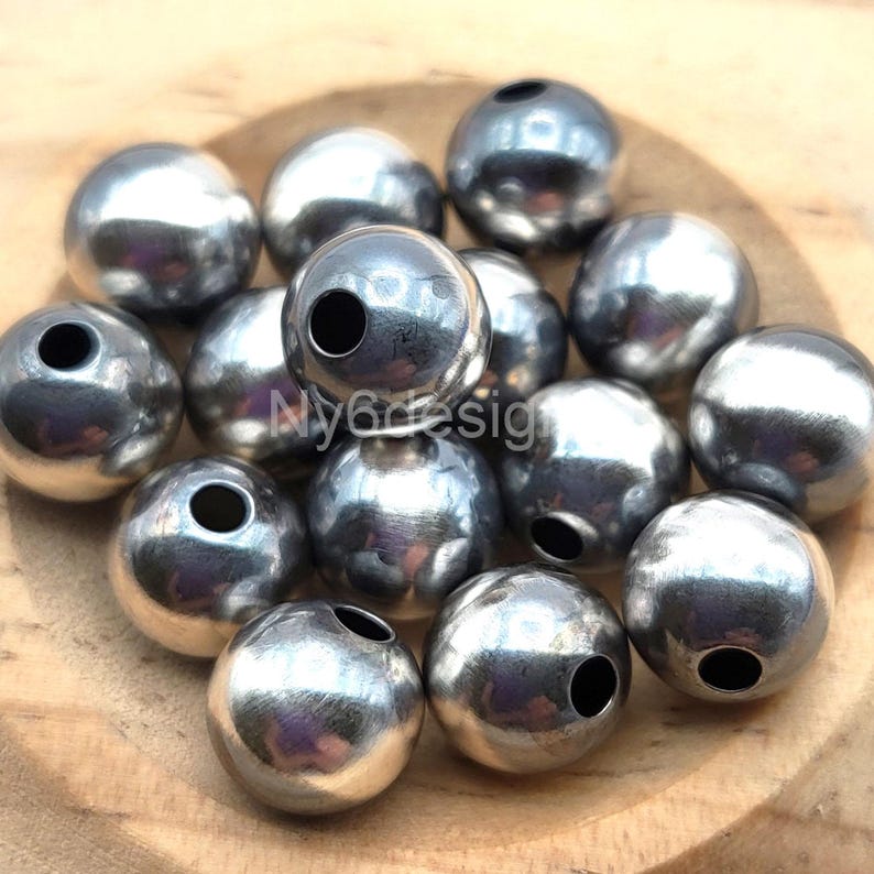 May include: A collection of shiny, silver-colored metal beads. Each bead is spherical with a hole drilled through the center. The beads are arranged on a wooden surface. The text "Ny6design" is visible on the image.