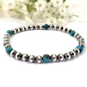May include: A silver beaded bracelet with turquoise accent beads. The bracelet features small, round, silver-colored beads interspersed with vibrant turquoise beads. The bracelet is displayed on a white surface with white flowers in the background.