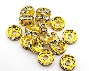 May include: A collection of gold-colored, donut-shaped beads with clear crystal accents. The beads have a shiny, metallic finish and are scattered on a white surface. These decorative beads are suitable for jewelry making and craft projects.