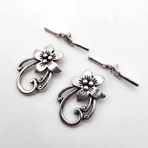 May include: Two silver-toned metal toggle clasps for jewelry making. Each set consists of a floral-shaped loop component with five petals and three central dots, paired with a straight bar component featuring a central loop attachment point.