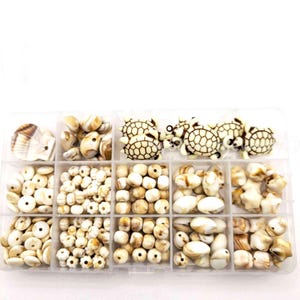 May include: A clear plastic organizer box containing various off-white and tan beads. The collection includes turtle-shaped beads, shell-shaped beads, star-shaped beads, and numerous round, oval, and disc-shaped beads with marbled brown patterns.