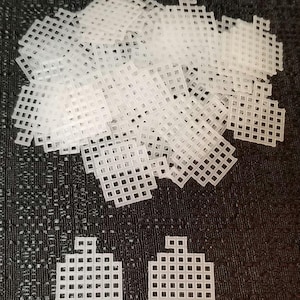 May include: A pile of white plastic canvas squares, with two individual squares in the bottom right corner. The squares are used for cross-stitch and other needlework projects.