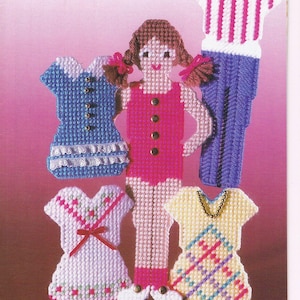 May include: A page from "Plastic Canvas World" magazine showing a magnetic doll and various outfits. The doll wears a pink dress and has brown pigtails. The outfits include a blue dress, a white dress with a red bow, and a yellow dress.