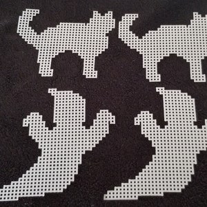 May include: Four white plastic canvas cutouts in the shapes of Halloween cats and ghosts. Two cats are side-facing with arched backs and tails. Two ghosts have outstretched arms. The cutouts are on a black background.