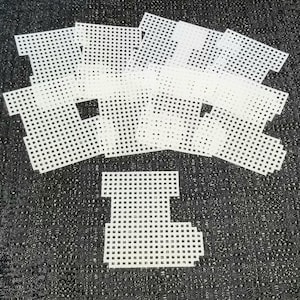 May include: White plastic canvas pieces in various shapes, including squares and a T-shape, are scattered on a dark, textured surface. The pieces have a grid-like pattern, suggesting their use in crafting or needlepoint projects.