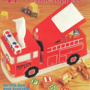 May include: A red plastic canvas tissue box cover shaped like a fire truck, with white accents and black wheels. The cover has a lift-up top and includes the text "MUNICIPAL FIRE DEPT". The image also shows alphabet blocks and the text "Seven fun and fanciful tissue box covers for the young at heart!"