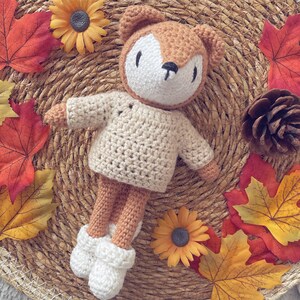 Puede incluir: Un juguete de zorro de crochet con un suéter de color crema, sentado en una estera de paja tejida con hojas de otoño y piñas.
