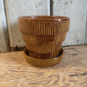 May include: A brown ceramic plant pot with a matching saucer, featuring a tiered design with a textured, basket-weave pattern on the exterior. The pot has a wide, open top and a tapered base, finished with a glossy glaze that highlights the details.