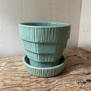 May include: A turquoise ceramic planter with a matching saucer. The planter has a tiered design with vertical grooves, and a crack is visible. The set is placed on a wooden surface.