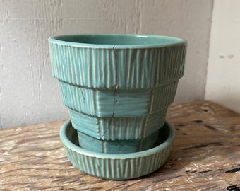 May include: A turquoise ceramic plant pot with a matching saucer. The pot has a tiered design with vertical grooves, and a crack is visible. The set is on a wooden surface.