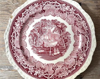 May include: A round, antique plate with a scalloped edge. The plate features a red and white design with a central landscape scene surrounded by a floral border. The plate's rim has a detailed, textured pattern.