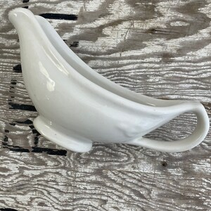 May include: A white ceramic gravy boat with a curved spout and a handle. The gravy boat has a smooth, glossy finish and sits on a small, round base. The design is classic and elegant, suitable for serving sauces and gravies.