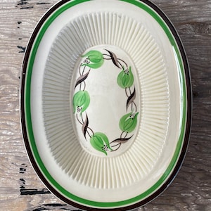 May include: An oval ceramic serving dish with a white background and a green and brown floral design. The dish has a raised rim and a scalloped edge.