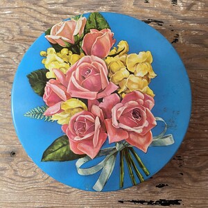 May include: A round, blue tin with a floral design. The lid features a bouquet of pink roses and yellow flowers tied with a light blue ribbon. Green leaves and stems complete the arrangement. The tin is likely vintage.