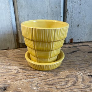 May include: A bright yellow ceramic plant pot with a matching saucer. The pot features a tiered, stepped design with vertical ribbed textures on each section, creating a basket-like appearance. The finish is glossy and uniform in color.