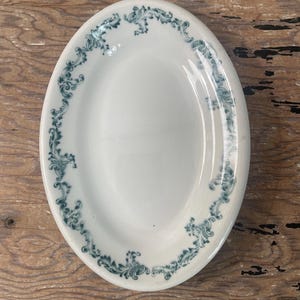 May include: An oval-shaped white ceramic platter featuring a decorative border of dark teal scrollwork patterns. The dish has a smooth, glossy finish and a plain center, with the intricate floral-inspired design framing the entire outer rim.