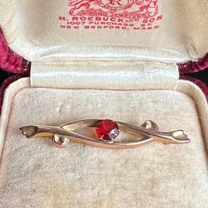 Puede incluir: Broche dorado con una gema roja central, exhibido en una caja de joyería de terciopelo rojo vintage abierta. El broche tiene un diseño delicado y curvado. El interior de la caja es de color crema.