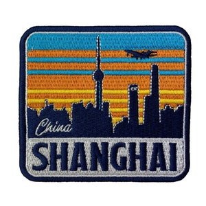 May include: An embroidered patch featuring a blue skyline of Shanghai, China. The patch has a sunset background with orange, yellow and blue stripes. The text "China" and "SHANGHAI" are embroidered in white.