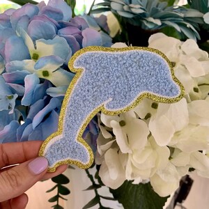 May include: A light blue felt dolphin with a gold glitter outline. The dolphin is a popular craft supply for making decorations and accessories.