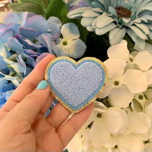May include: A heart-shaped patch featuring a textured light blue center, a smooth light blue border, and a shimmering gold glitter edge. The patch is layered with a soft, fuzzy material in the middle, creating a distinct contrast against the metallic gold trim.