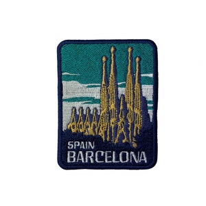 May include: A rectangular embroidered patch featuring a gold-colored depiction of the Sagrada Familia against a teal and white sky background. The bottom section is dark blue with the white embroidered text reading SPAIN BARCELONA.