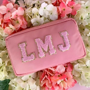 May include: A pale pink cosmetic bag featuring the embroidered initials "LMJ" in pink with gold trim. The bag is set against a backdrop of pink and white hydrangeas, offering a delicate, floral presentation.