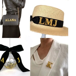 May include: A black crossbody bag with the name ALANA in gold, a straw fedora with a black ribbon and gold LMJ initials, a clear ornament with gold flakes and a black velvet bow with gold A and J, and a white blazer with a gold K initial pin.